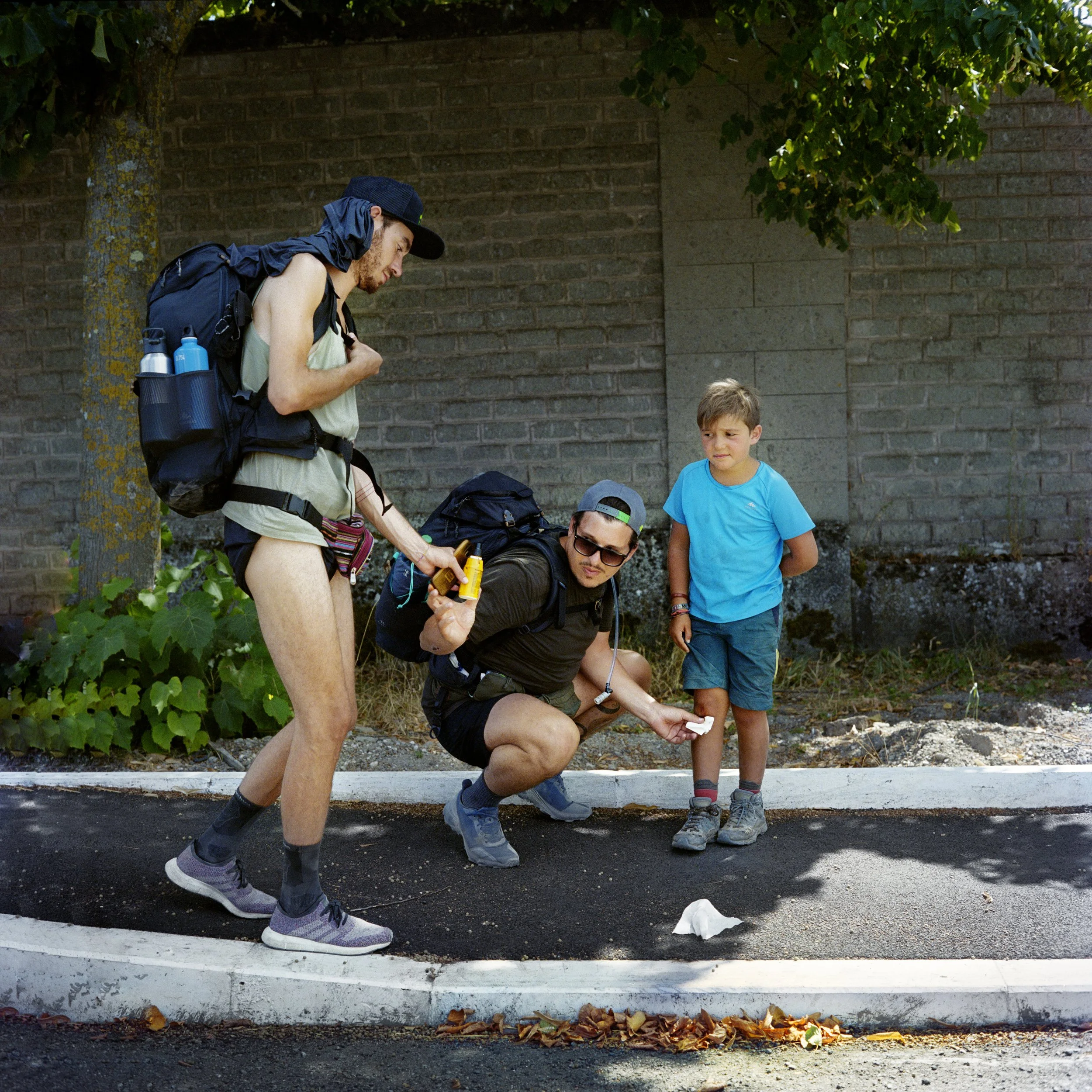 Two adults and a child pose on a sidewalk, apparent as if on an excursion or hike, with backpacks and casual clothes, under a tree, with a wall in the background.