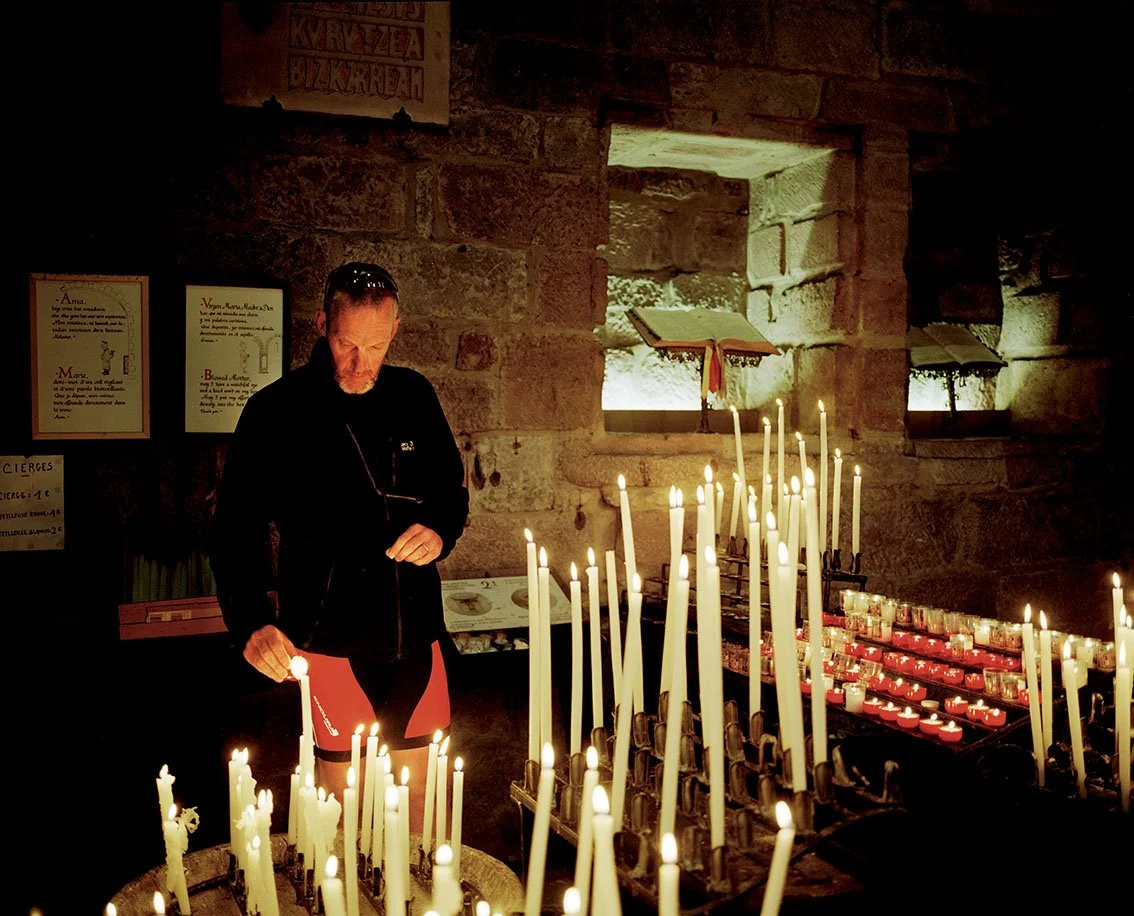 A man in a church lights a candle on a candelabra filled with lit candles, with stone walls and lit windows in the background.