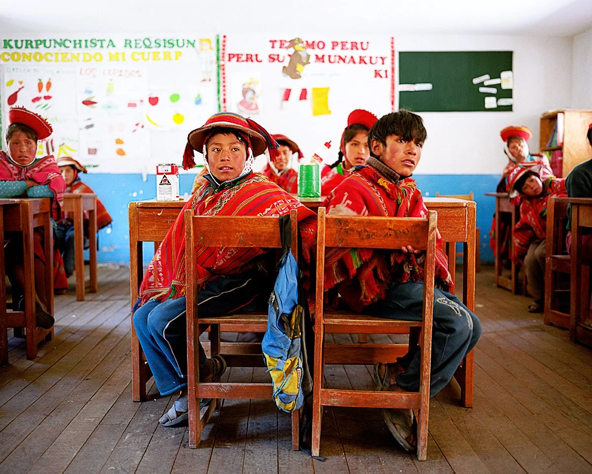 Children sitting in a classroom, wearing colorful traditional clothes and hats. The board behind them features drawings and texts, indicating that they are probably in a school in Peru.