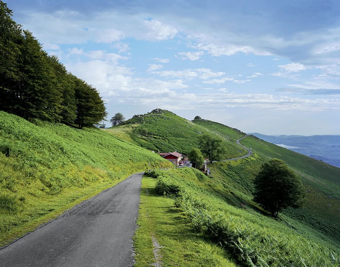 Asphalt road winding through a lush green hill with a few trees, wooden houses and a clear view of other mountains in the distance, under a partly cloudy sky.