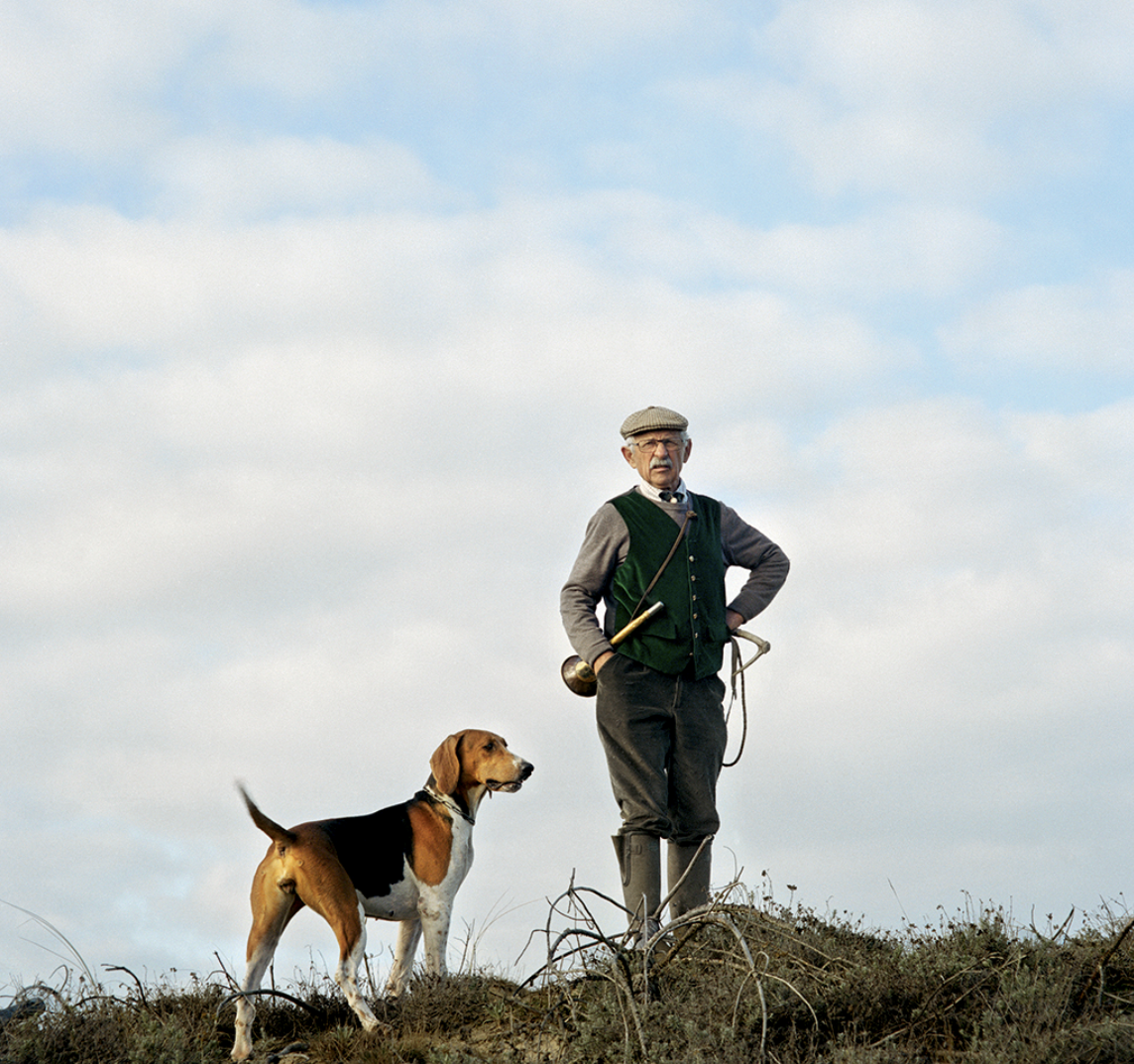 Un homme âgé avec un chien de chasse sur un terrain herbeux sous un ciel principalement dégagé.