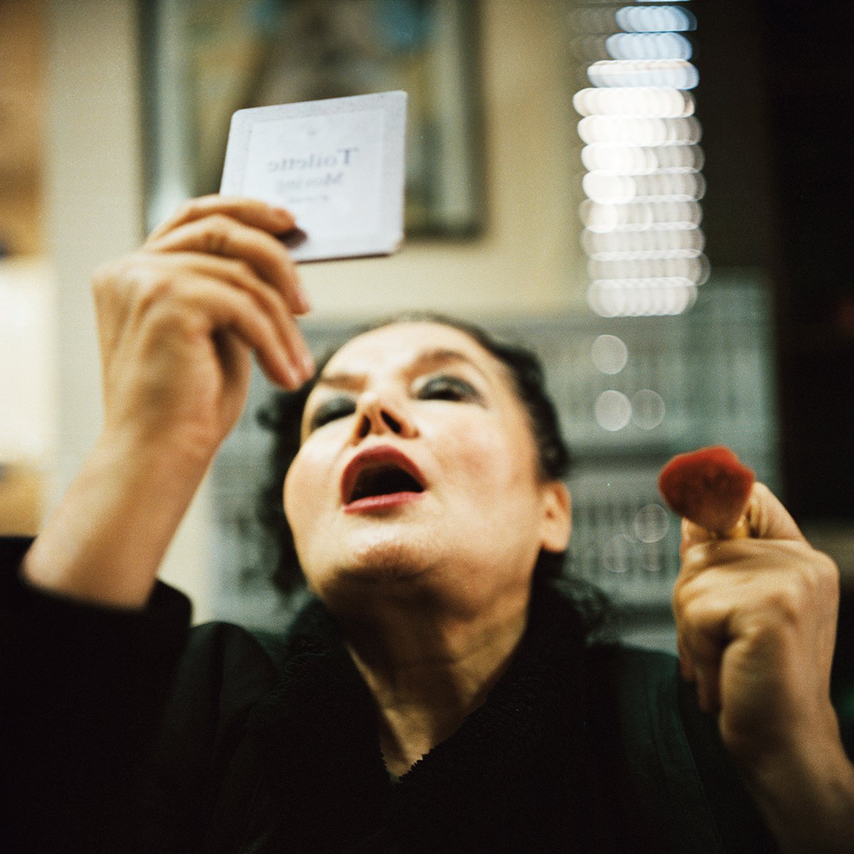Une femme avec expression provocante regarde un miroir, tenant un objet rouge dans la main droite et un ticket dans la main gauche.