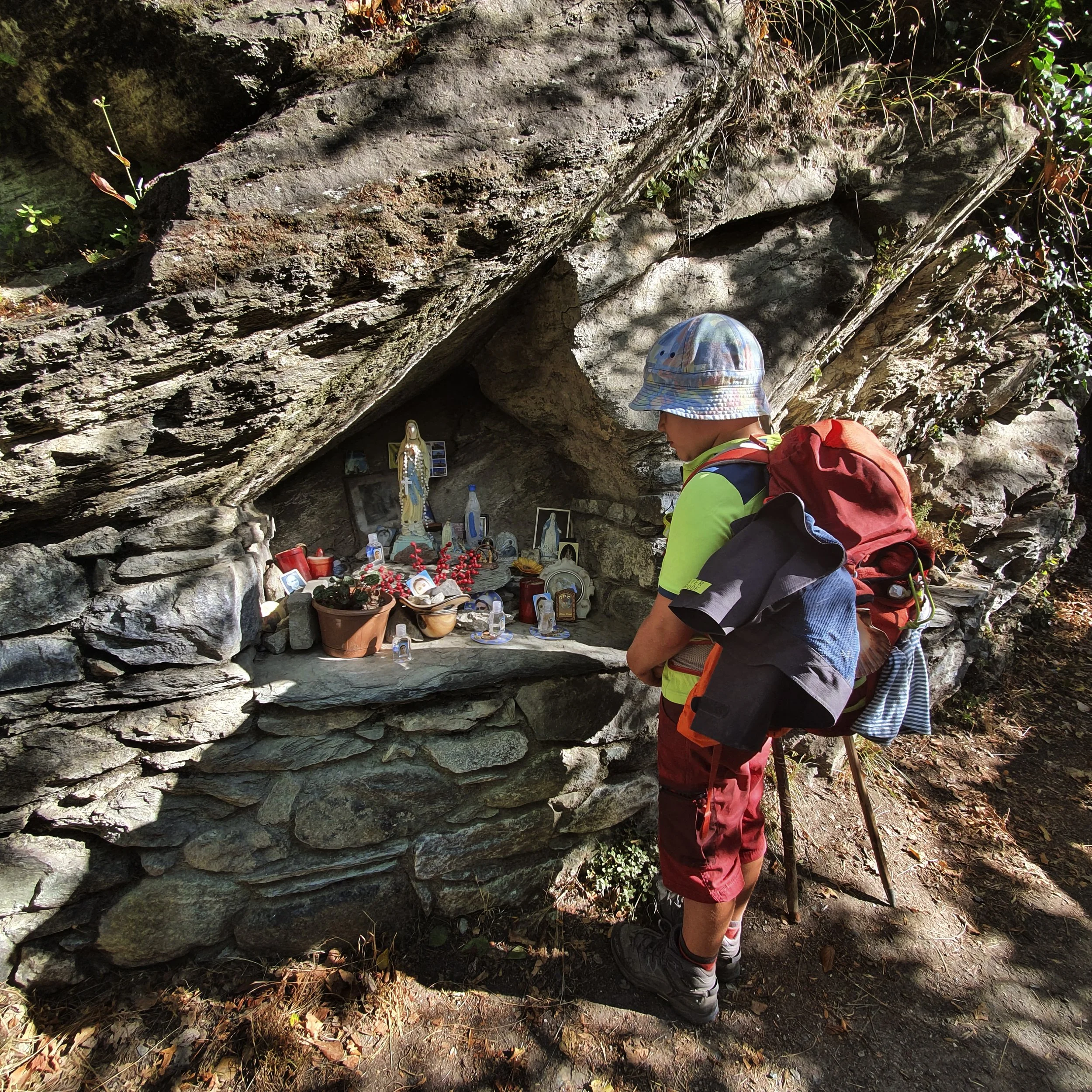 A child with a backpack and a hat observes a small chapel or altar dedicated to the Virgin Mary, with various statues, photos, candles and flowers placed in a rock niche in the open air.