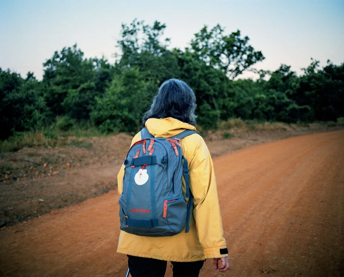 Person seen from the back wearing a blue backpack and yellow jacket, walking along a country lane surrounded by green trees, at dusk.