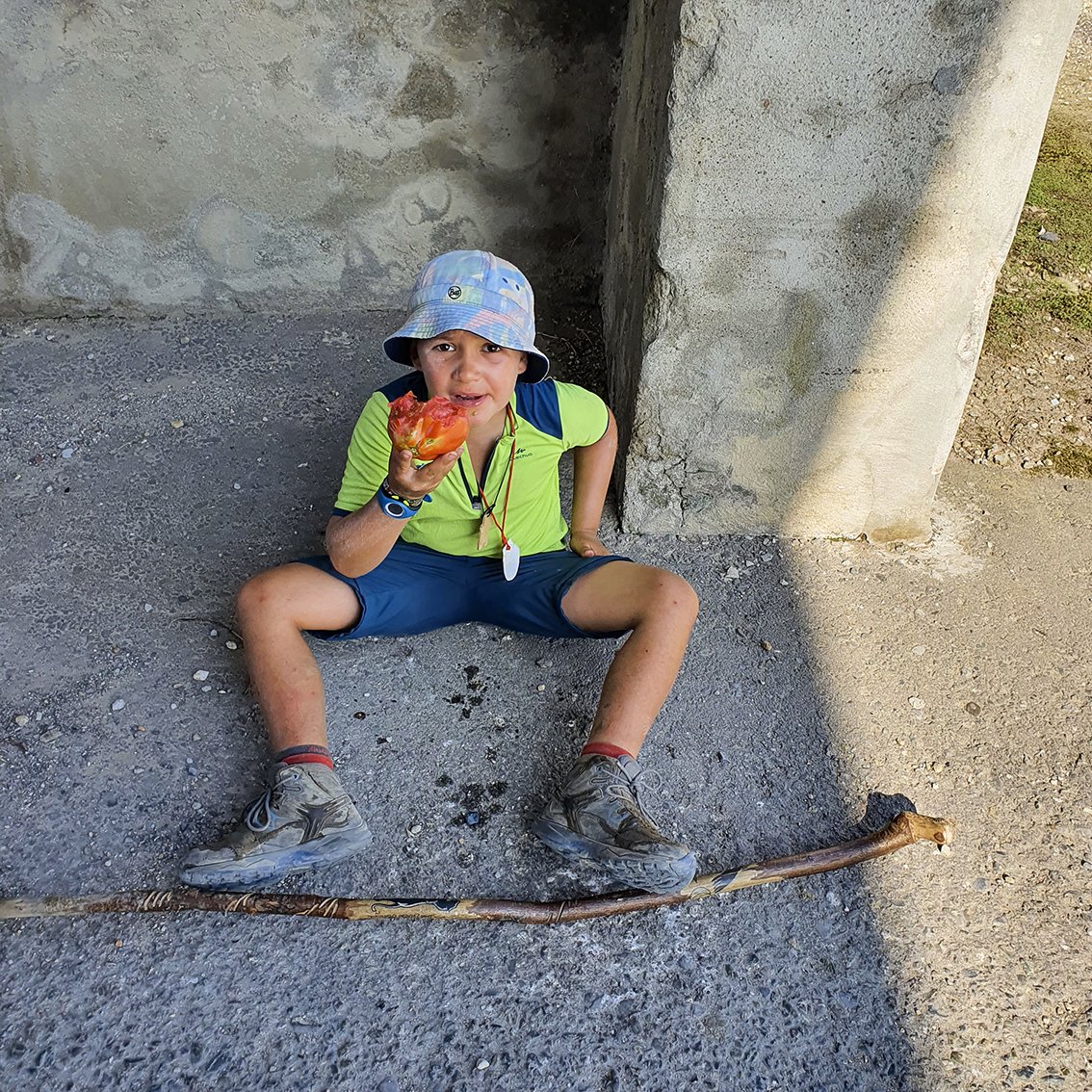 A boy sits on the ground, wearing a canvas hat, a green T-shirt and blue shorts, eating a ripe tomato by hand. He's leaning against a concrete wall, with a wooden cane resting in front of him.