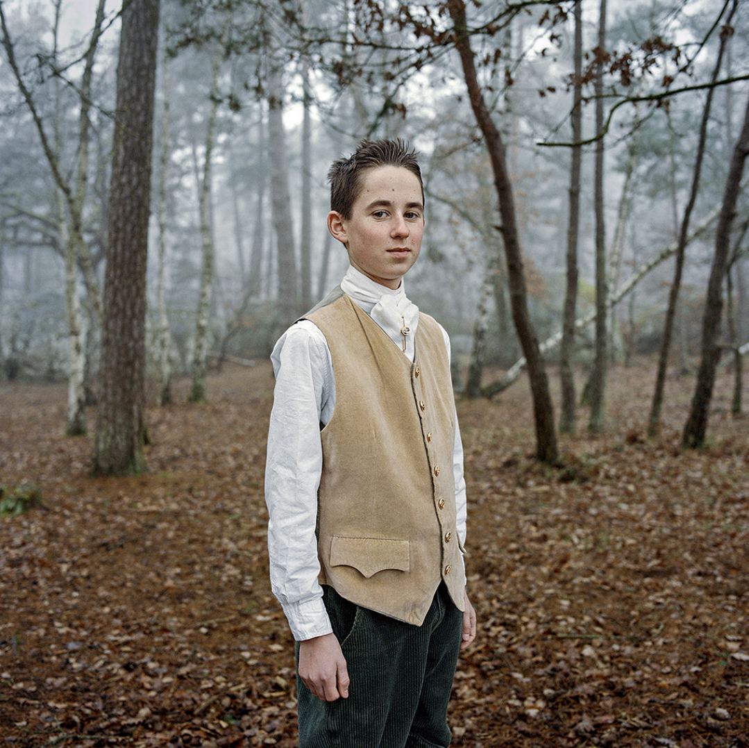Jeune homme en vêtements vintage, debout dans une forêt brumeuse automnale, portant une veste beige et une chemise blanche.