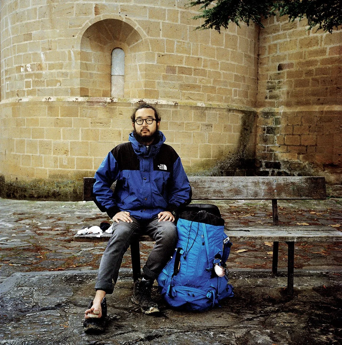 A man sitting on a stone bench in front of a brick wall. He's wearing a blue and black jacket, glasses, and has a large blue satchel beside him. He has a beard, long hair tied back, and a black beard.