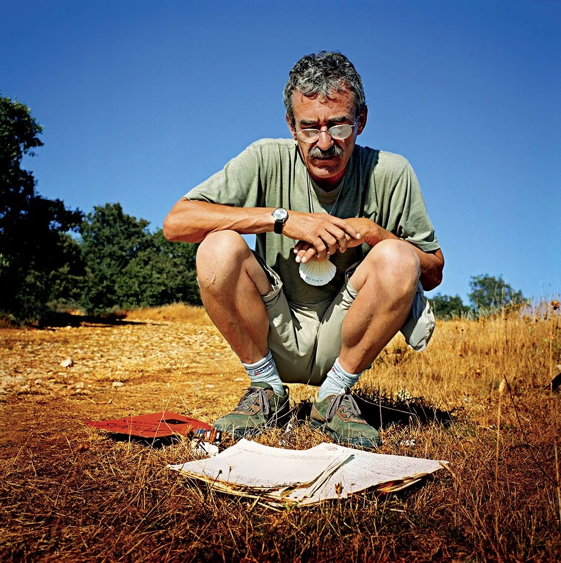 An elderly man, wearing sunglasses and hiking gear, crouches in a dry field. There are papers and a notebook in front of him, under a clear blue sky.