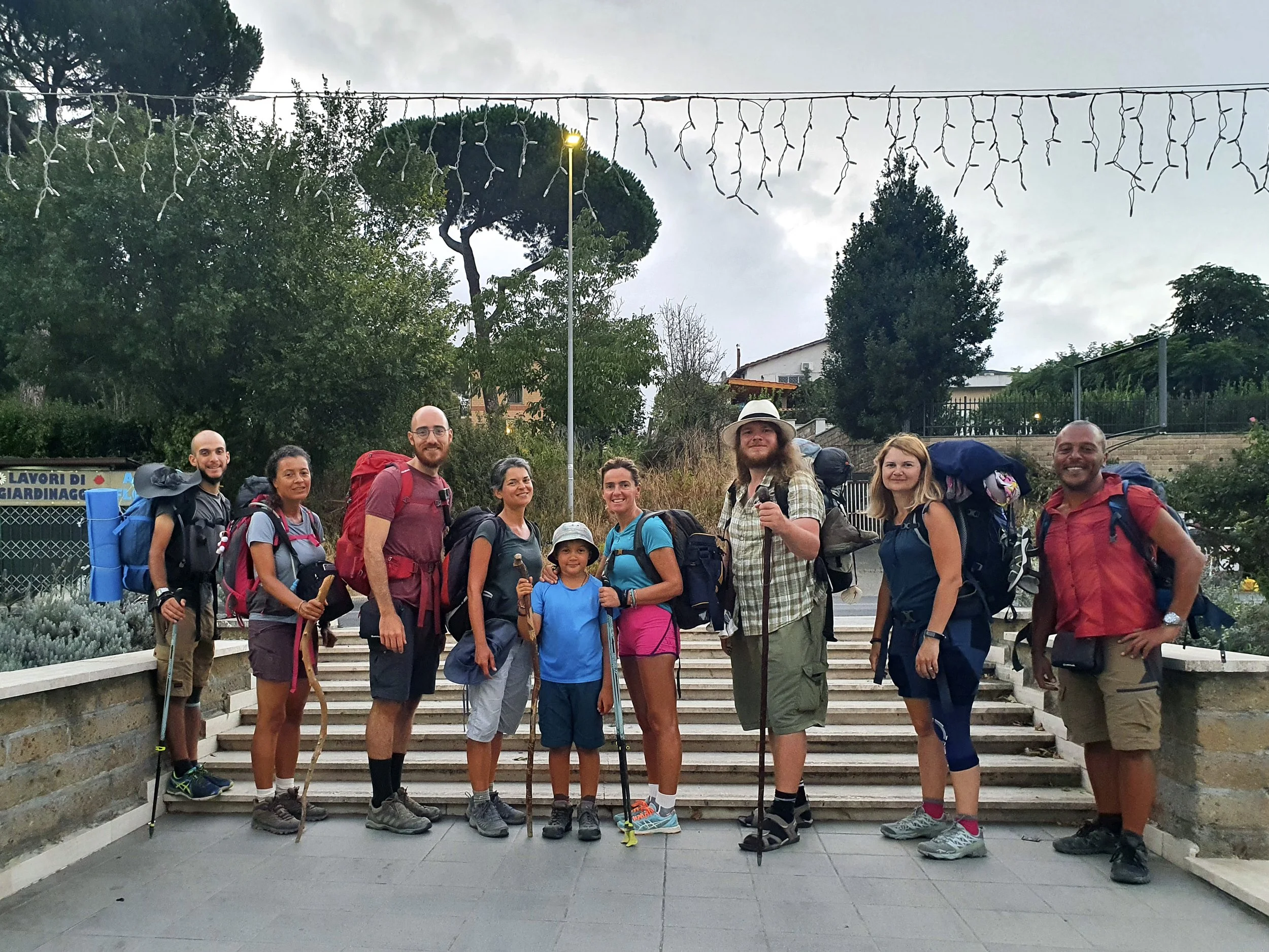 Group of nine hikers with backpacks, standing on outdoor steps, with trees and a cloudy sky in the background.