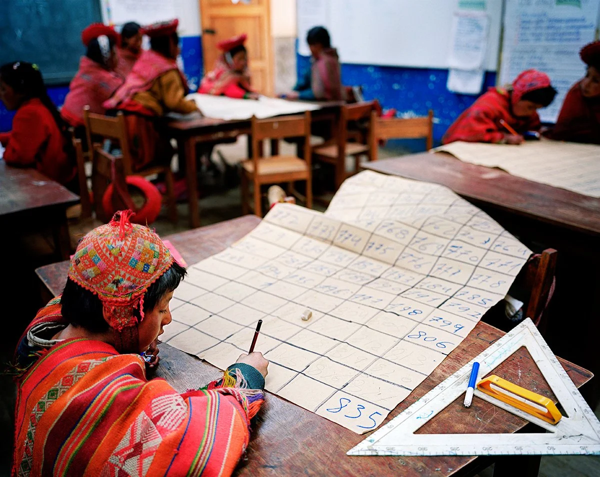 A child in colorful traditional clothing works on a large flipchart with numbers, in a classroom. Other children in traditional dress sit at tables in the background.