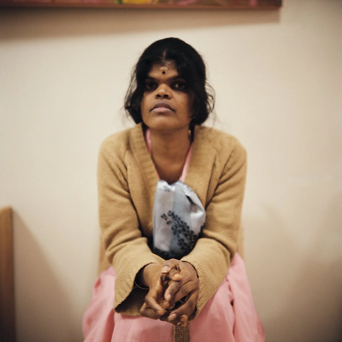 Femme assise dans une pièce, affichant une expression sérieuse, portant un pull beige, une robe rose, avec un fond neutre.