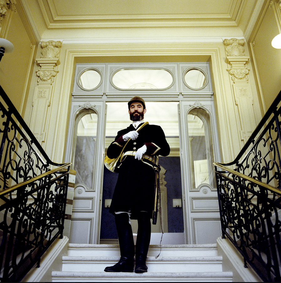 A man in historic uniform, complete with hat, white gloves and gold cord, stands on a marble staircase with wrought-iron banisters, in an elegant interior with richly decorated architectural details.
