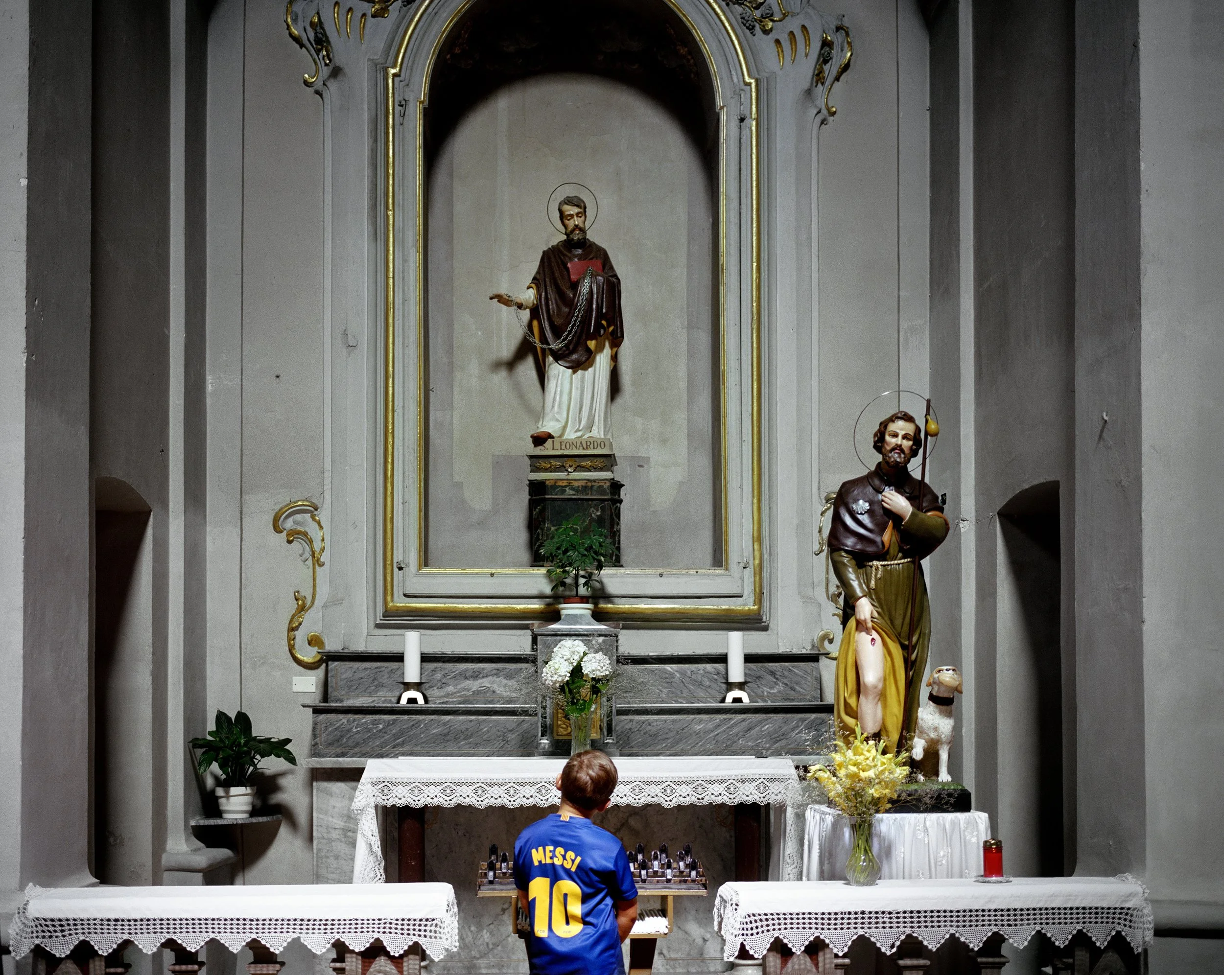 A boy looking at a small chess table in front of an altar in a church, with a statue of Jesus and another of a saint, flowers and candles.