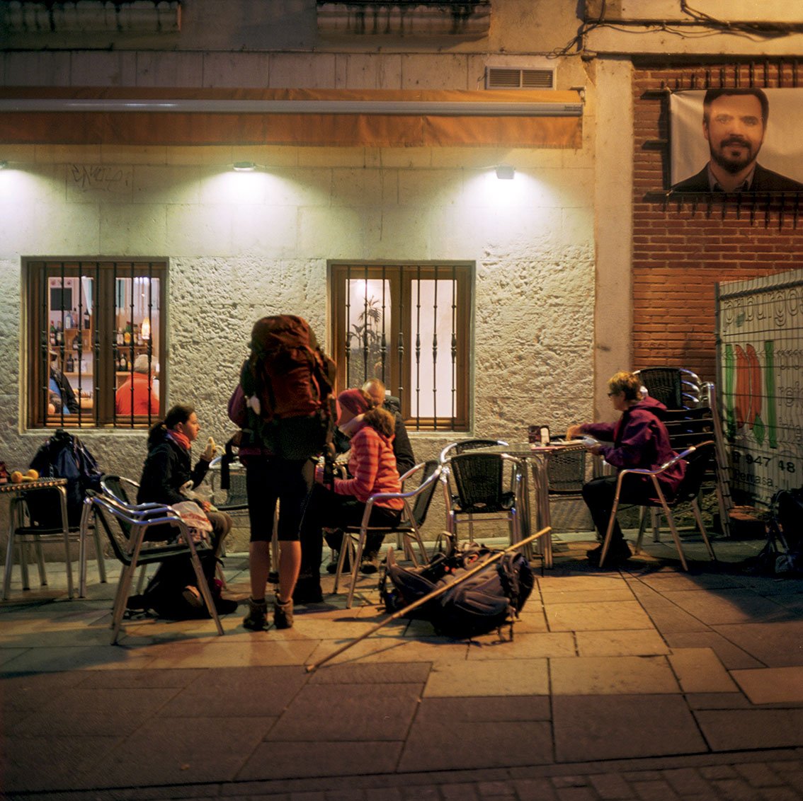 Group of people sitting on a café terrace in the evening, chatting and eating, with a man sitting alone on the right, with bags and a stick on the ground, in front of a building with windows, a portrait poster, and a brick and plaster wall.