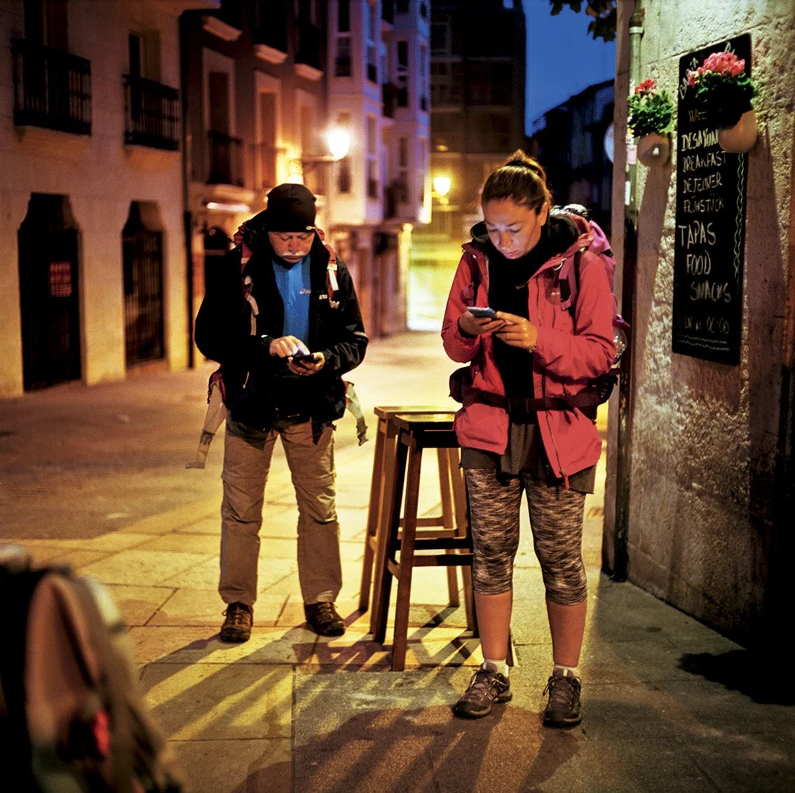 Two people dressed in hiking clothes, looking at their phones, standing on a sidewalk at night, next to a black sign with a list of dishes, on a lit urban street.