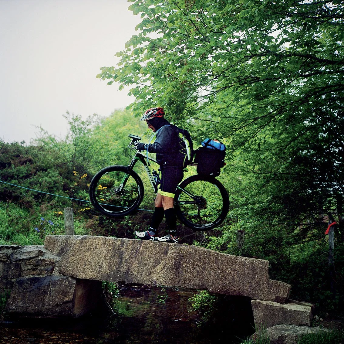 A cyclist wearing a helmet and carrying a rucksack, lifting his bike over a stone bridge in a green forest.