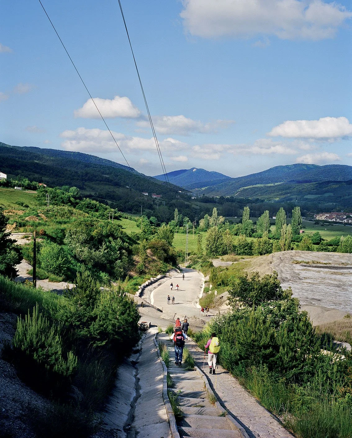 Mountain landscape with a hiking trail and people walking, surrounded by green vegetation, blue sky with a few clouds.