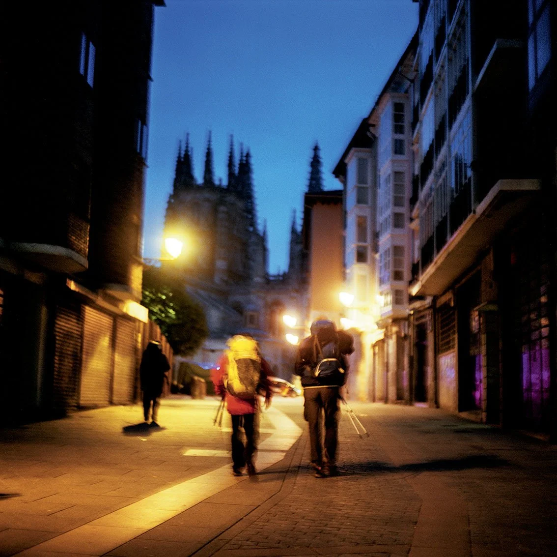 Urban street at night with people walking, modern and old buildings, Gothic church in background, street lamps illuminating the scene.