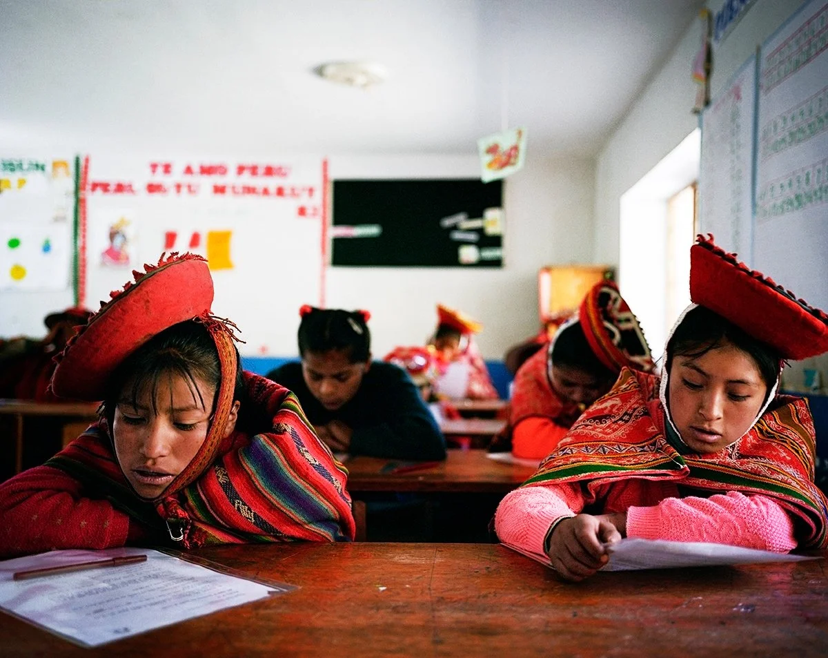 Mexican children in traditional uniforms, wearing red hats, sit at their desks in class, concentrating on their work.