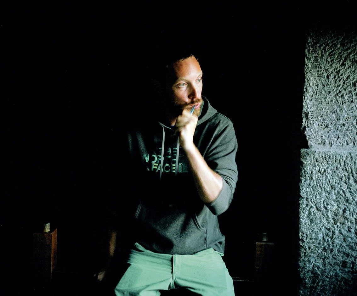 Young man with red hair and beard, wearing a dark gray hoodie, standing next to a stone wall in a dark room. He looks pensively to the left, with his hand resting under his chin.