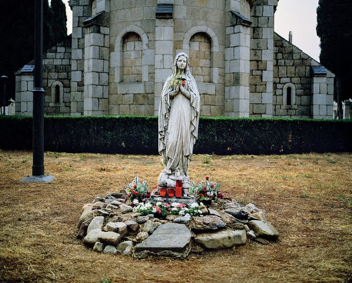Statue of the Virgin Mary surrounded by flowers and candles, in front of a stone church.