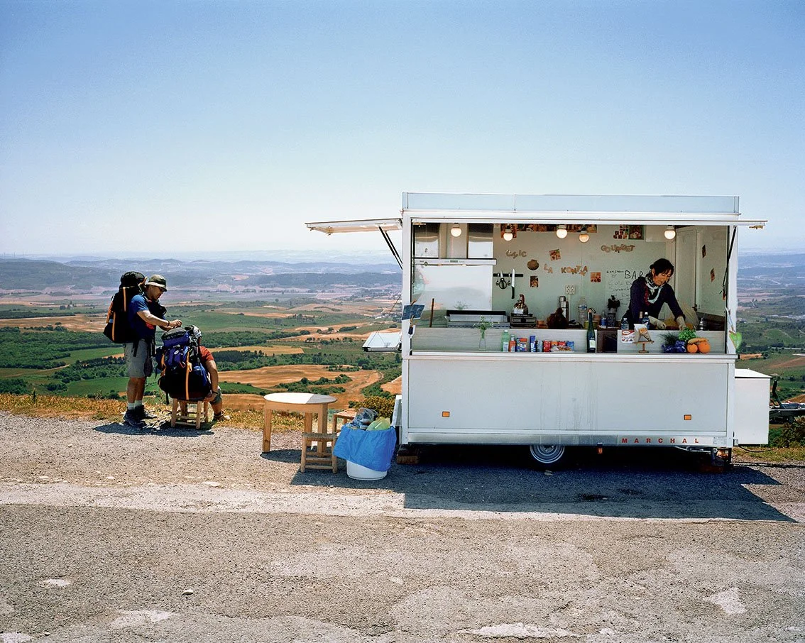 A white food truck parked on the side of a road with a panoramic view of the countryside, a woman preparing something inside, and three people with backpacks standing next to it.