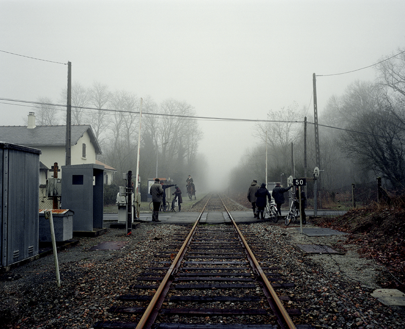 Vue d'une gare routière avec des barrières et des voies de chemin de fer dans un brouillard, avec plusieurs personnes, dont des cyclistes et un homme à cheval, attendant ou traversant la voie.