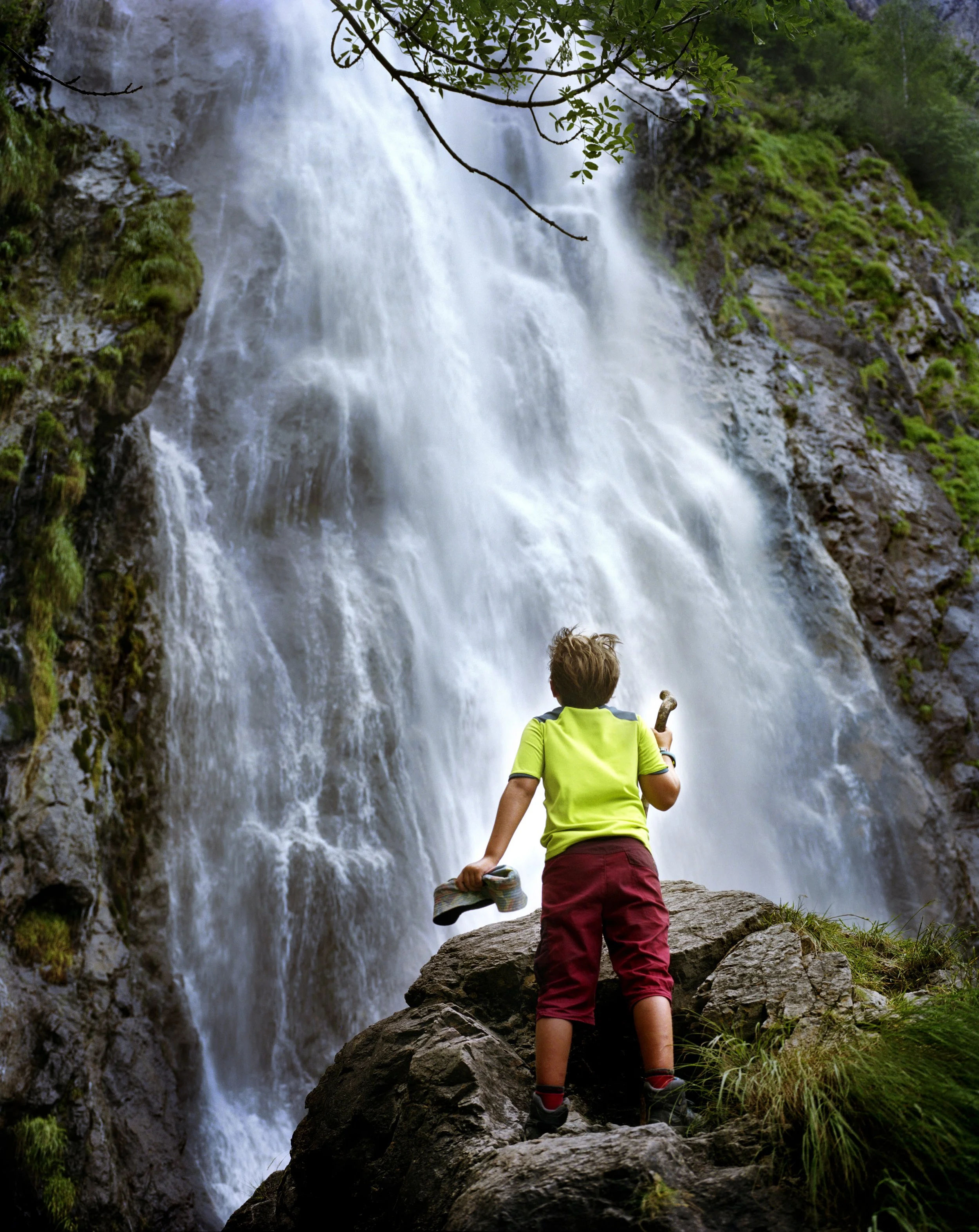 Young boy in green T-shirt, red pants, standing on a rock, looking at a waterfall in the middle of nowhere.