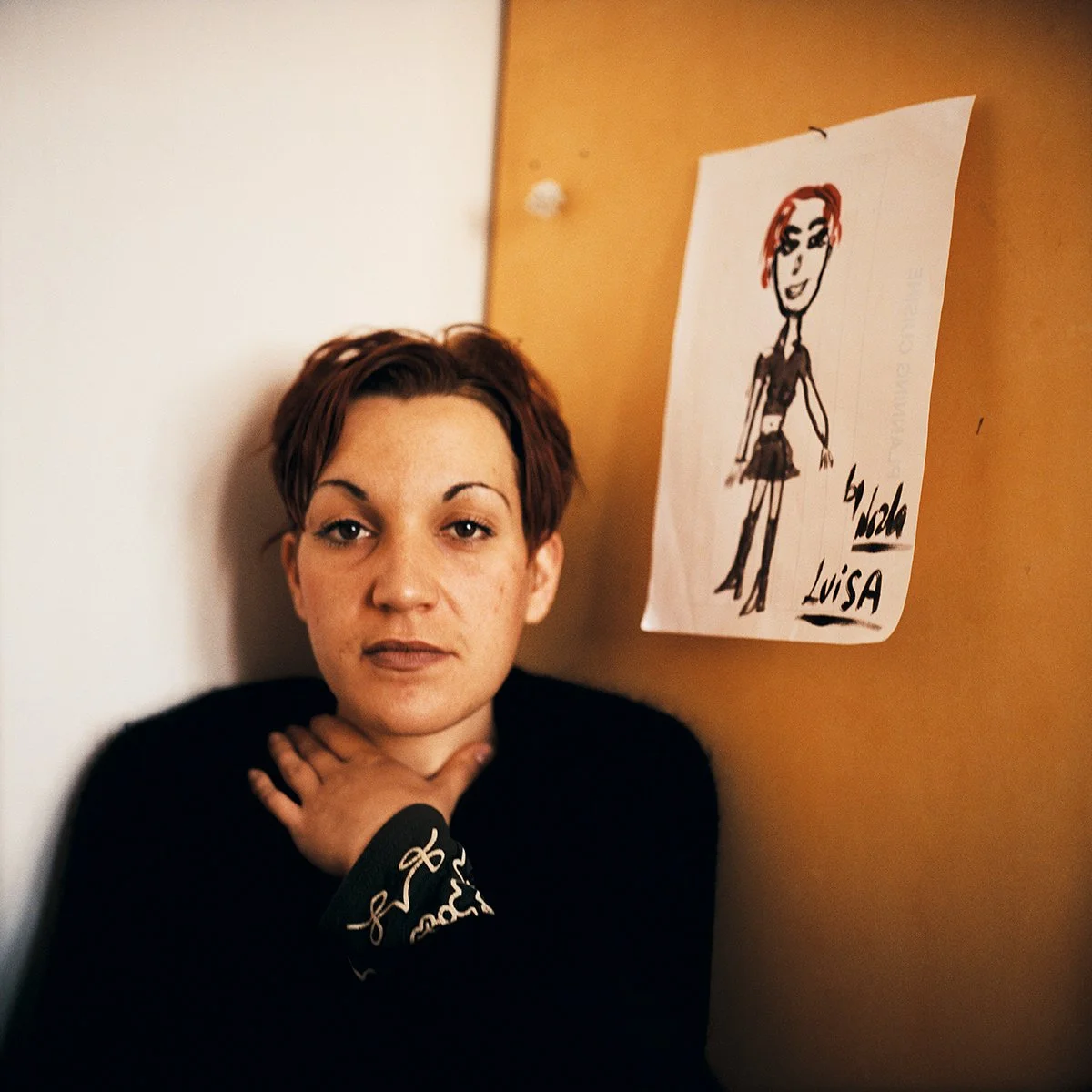 Jeune femme aux cheveux courts, portant un vêtement noir, posant près d'un mur à côté d'un dessin d'une femme avec des cheveux roux et un short noir, avec le texte "Chloé" et "Lisa" à côté.