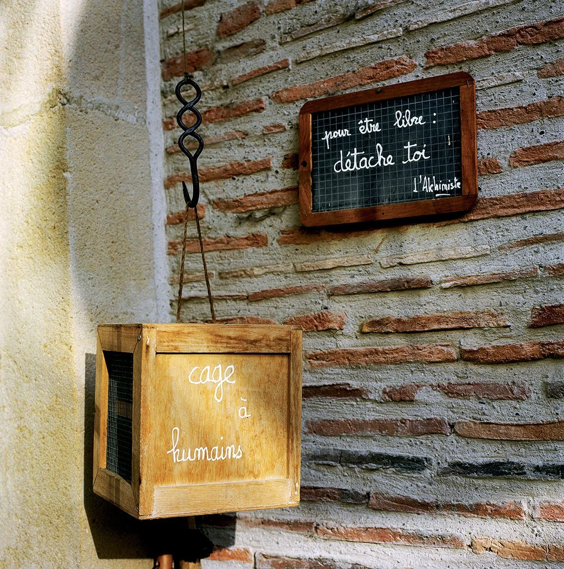 A wooden box with "human cage" written in white, hung on a brick wall. Next to it, a blackboard with a white inscription: "to be free: untie yourself, L'Alchimiste".