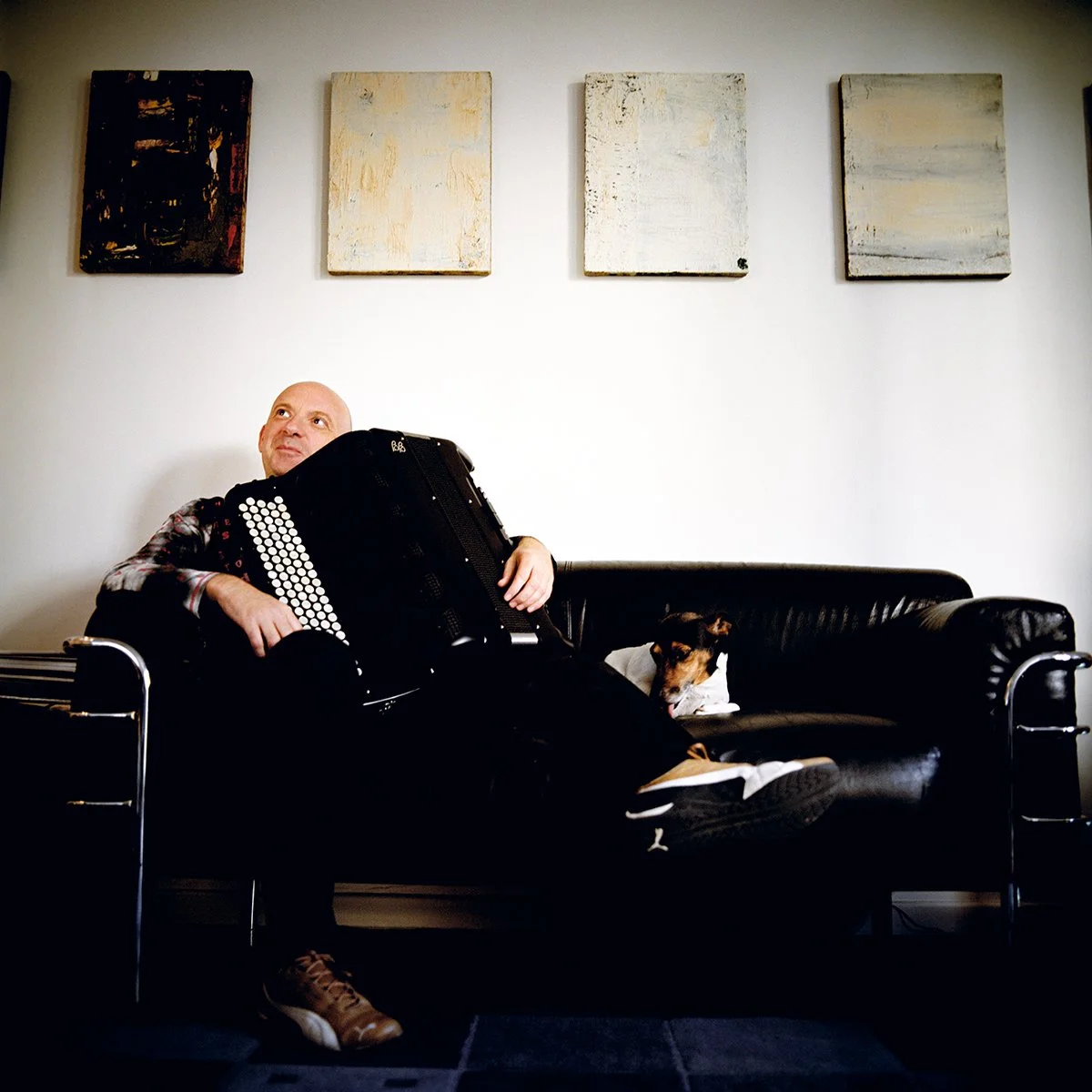 A man sits on a black sofa with a small dog lying next to him, a woman plays the accordion on her lap, in a room with a white wall and four abstract paintings hanging.