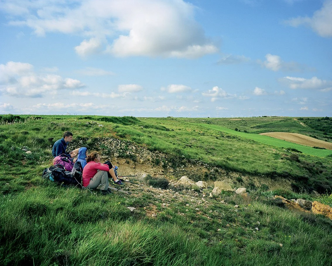 Three people on a hike stop near a damaged area in a verdant rural landscape under partly cloudy skies.