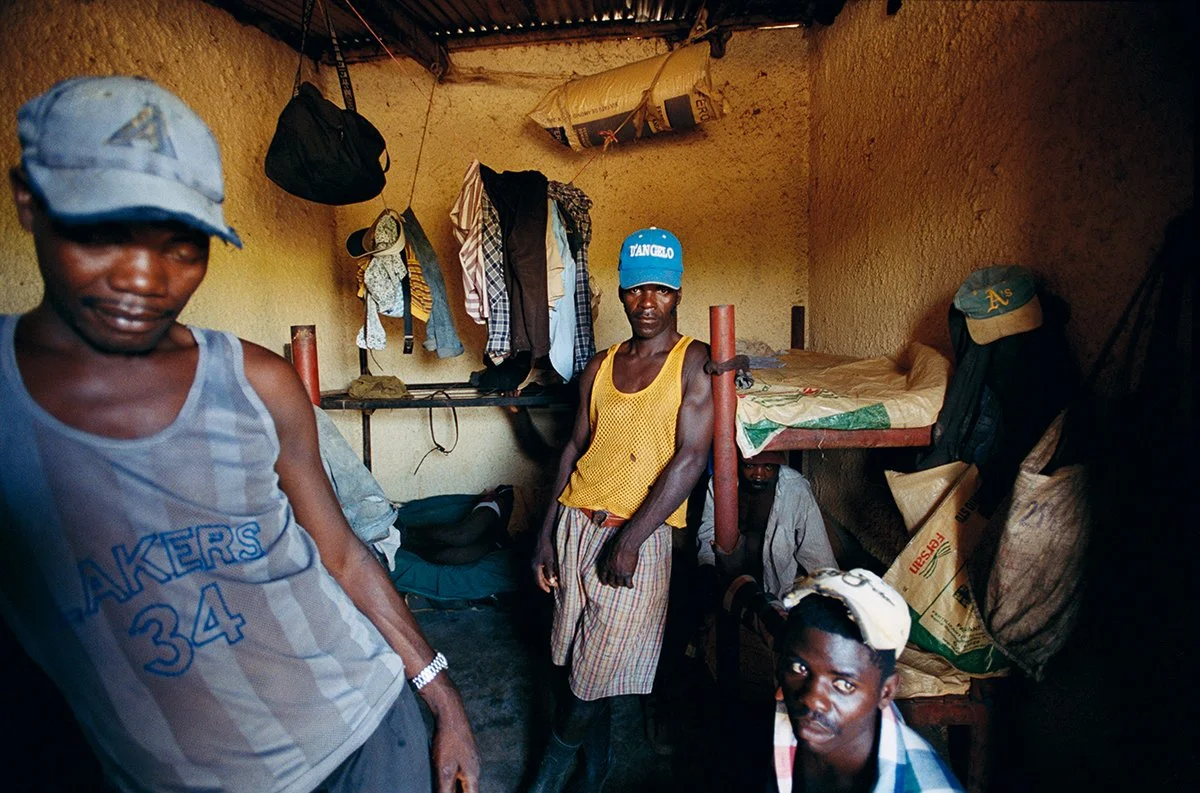 Five men in a small room with mud walls, hanging clothes, a bed and bags. One wears a blue cap, another a plaid shirt, and they all have varied expressions.
