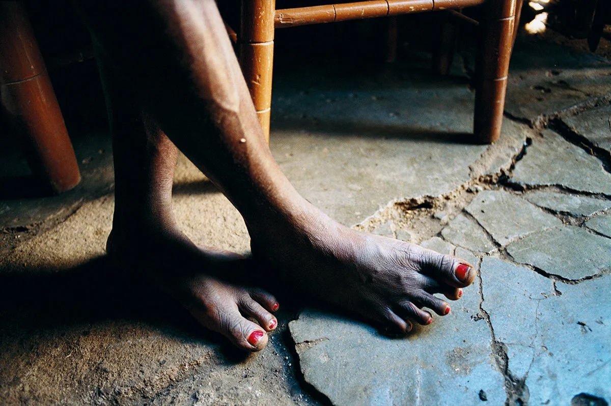 The feet of a person with nails painted red, placed on a stone and earth floor, under a wooden chair.