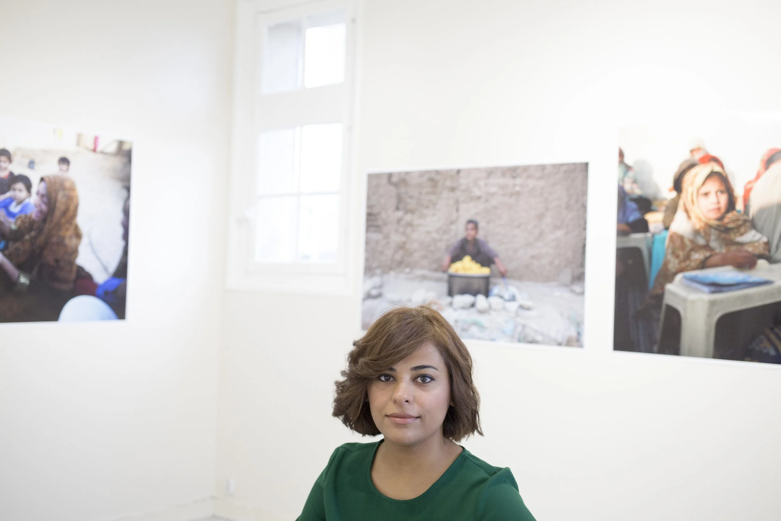 Young woman with curly brown hair, wearing a green top, sitting in an art gallery with photographs hanging on the wall.