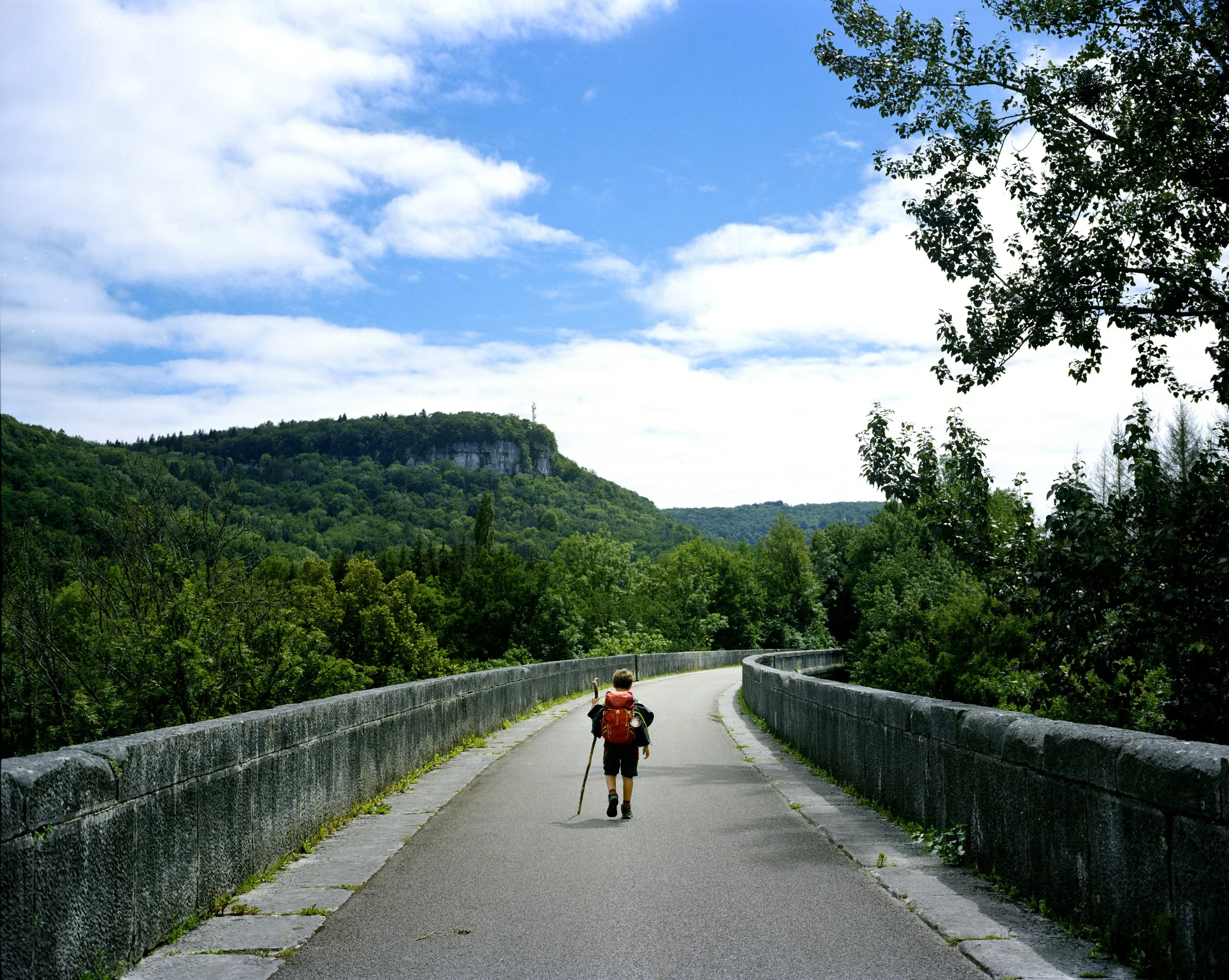 A young person with a backpack walking on a stone bridge, surrounded by lush vegetation and a blue sky with a few clouds.