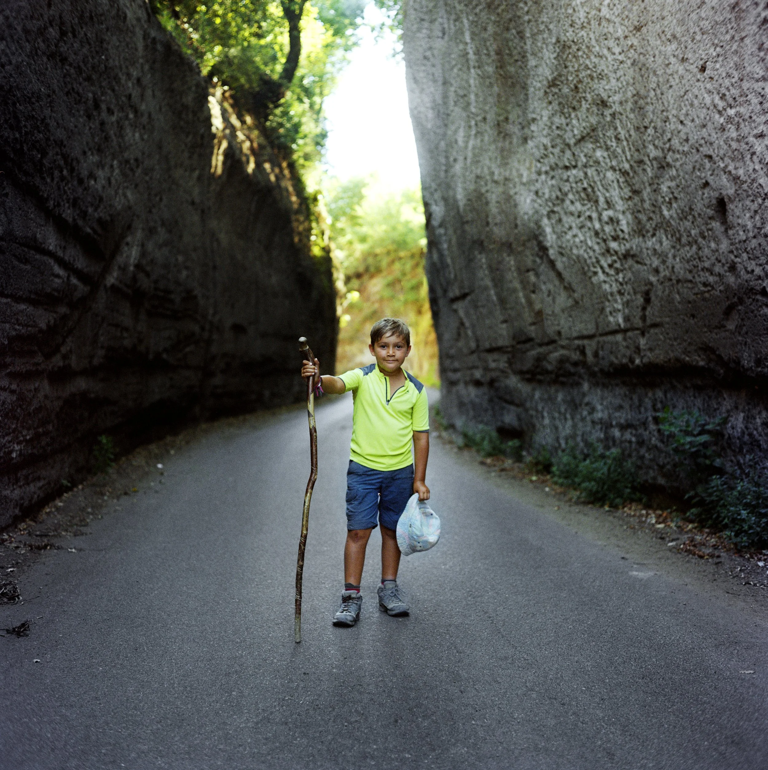 A boy standing on a path surrounded by high rock faces, holding a stick and a plastic bag, smiling at the camera.
