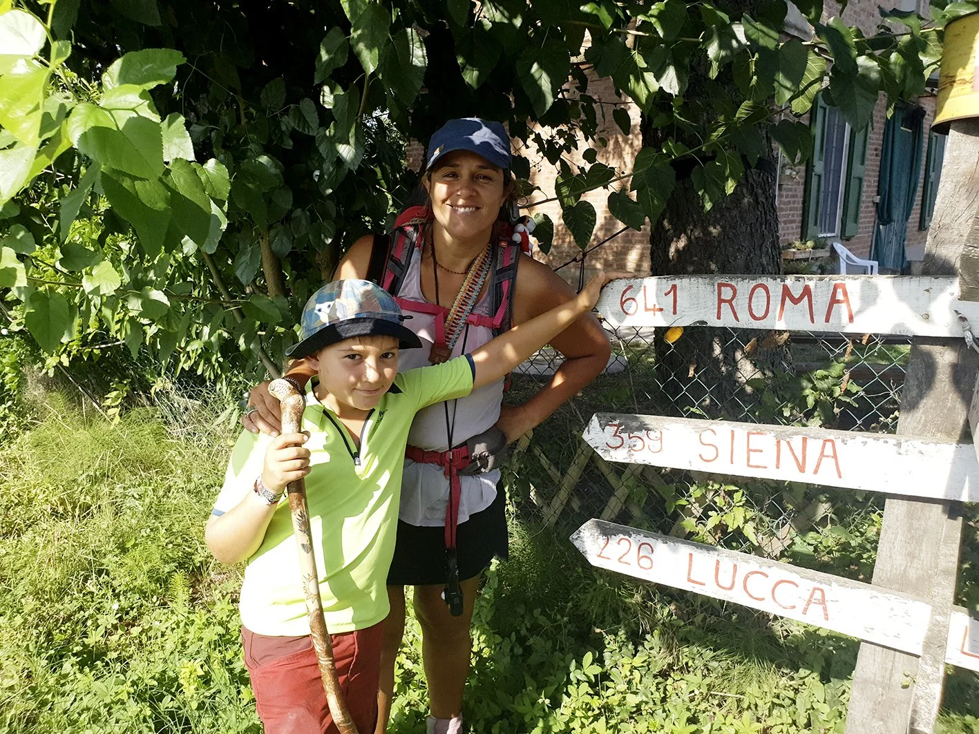 A woman and child on a hike, pointing a direction on a wooden sign with the names of towns, in verdant Italian surroundings.