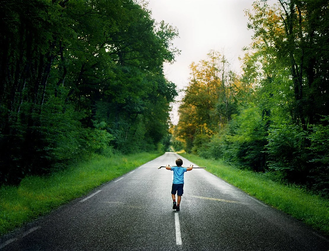 A child walks alone on a hilarious road on the edge of a verdant forest, with trees of green and yellow leaves, under a cloudy sky.