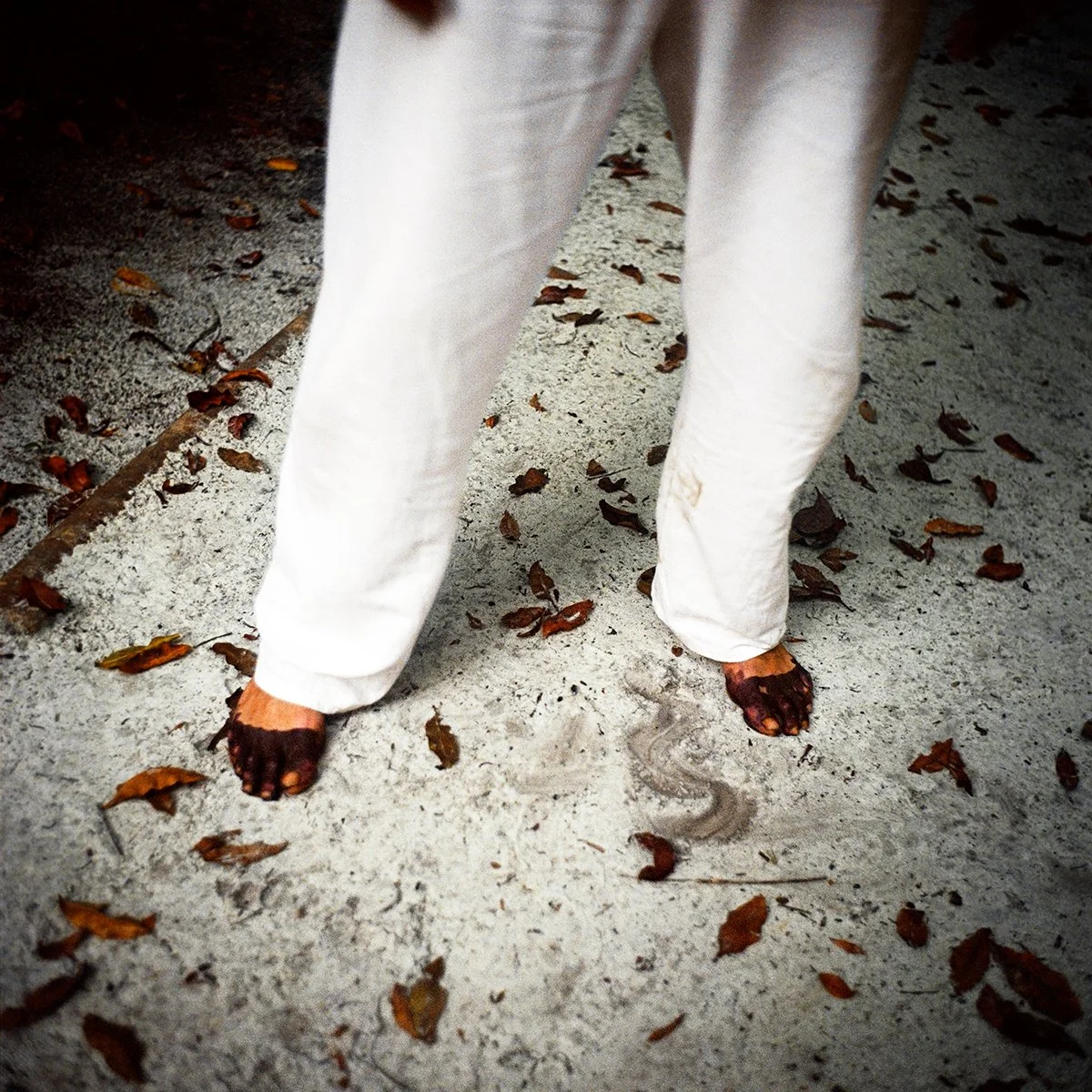 You see a person dressed in white pants, barefoot, walking on a concrete floor strewn with dead leaves, with part of his leg visible.