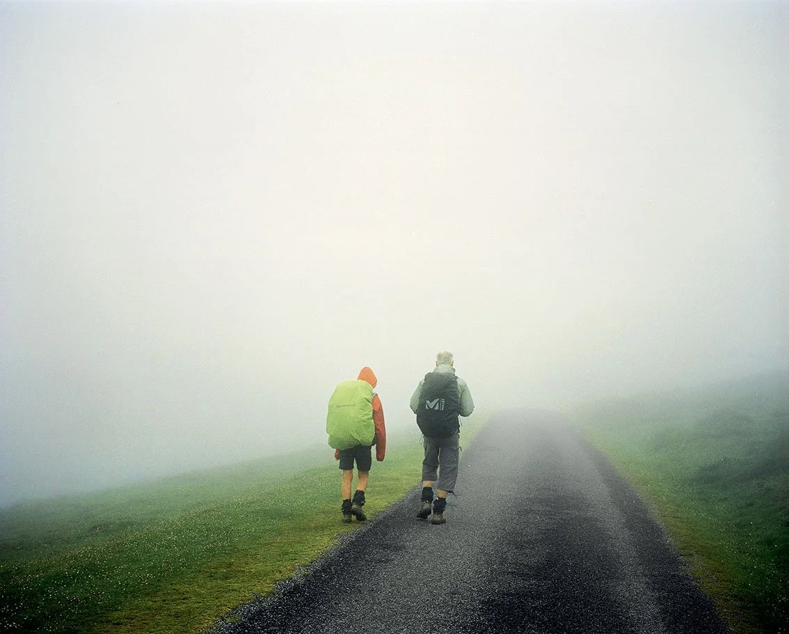 Two hikers walking along a foggy path, carrying backpacks. The scene is shrouded in dense fog.