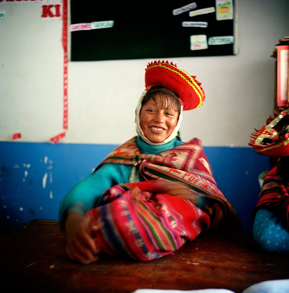 A smiling girl wearing a colorful traditional garment with a red hat in a room.