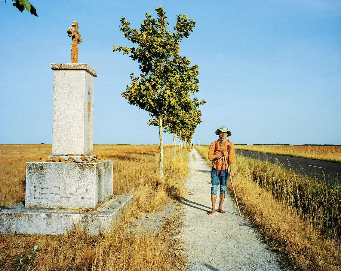 A woman walking along a rural path with a walking stick, wearing a hat, orange shirt and backpack, next to a stone cross in a dry field and a clear blue sky.