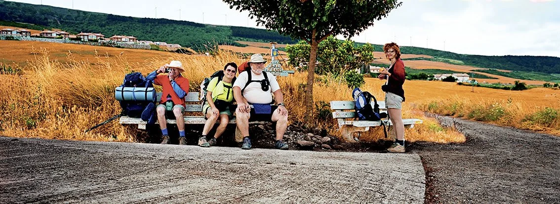 Four hikers sitting on a bench and a woman standing next to them in a field with hills in the background, carrying backpacks.