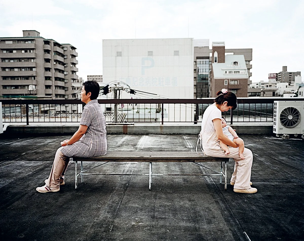 Une femme avec un bébé assise sur un banc sur le toit d'un bâtiment, avec un homme assis de l'autre côté, regardant dans des directions opposées, dans un environnement urbain.