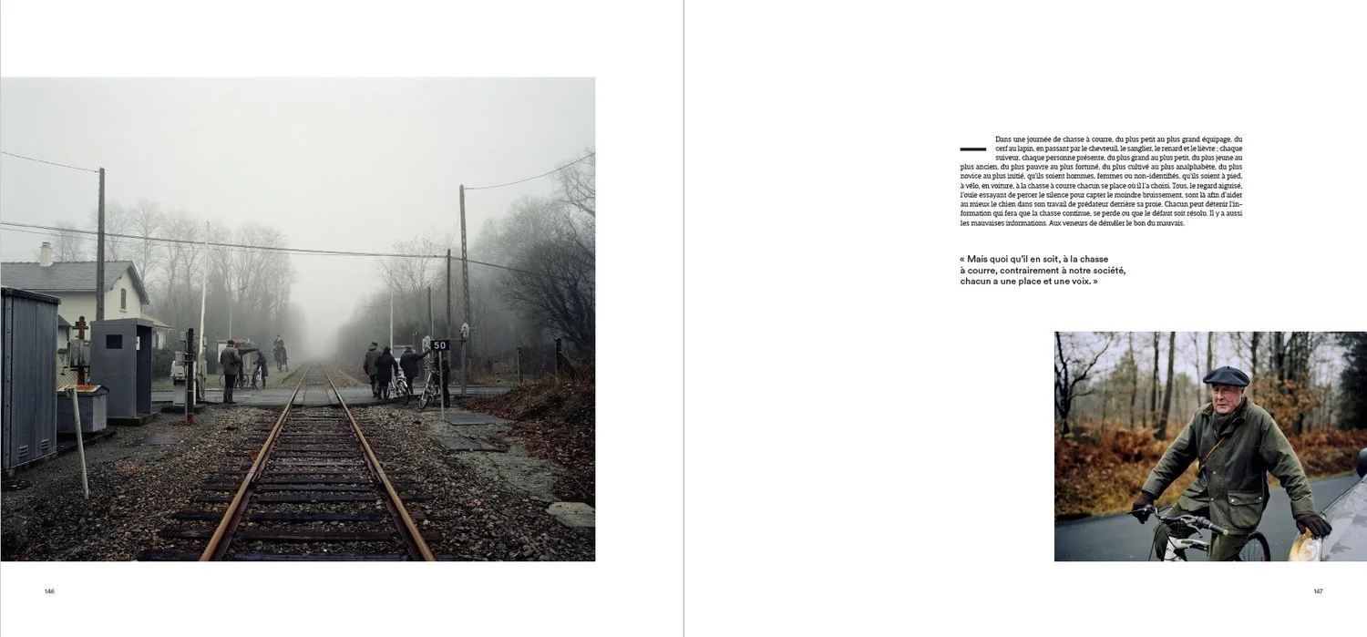 Vue de nuit sur une voie ferrée avec des passagers attendant, brouillard, arbres sans feuilles et maisons en arrière-plan.