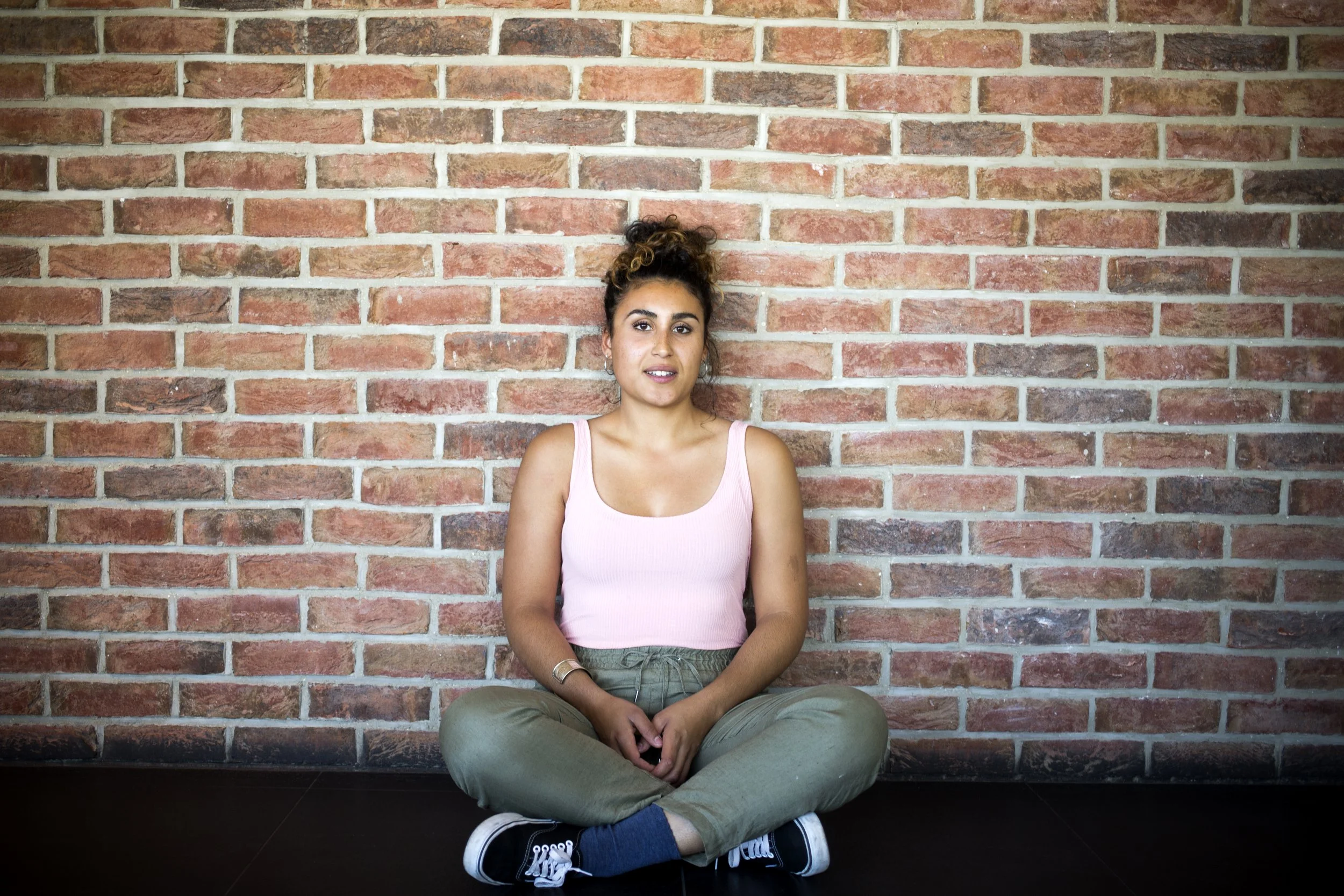 A young woman is growing, sitting on the ground against a red brick wall, wearing a pink top, beige pants and black sneakers.