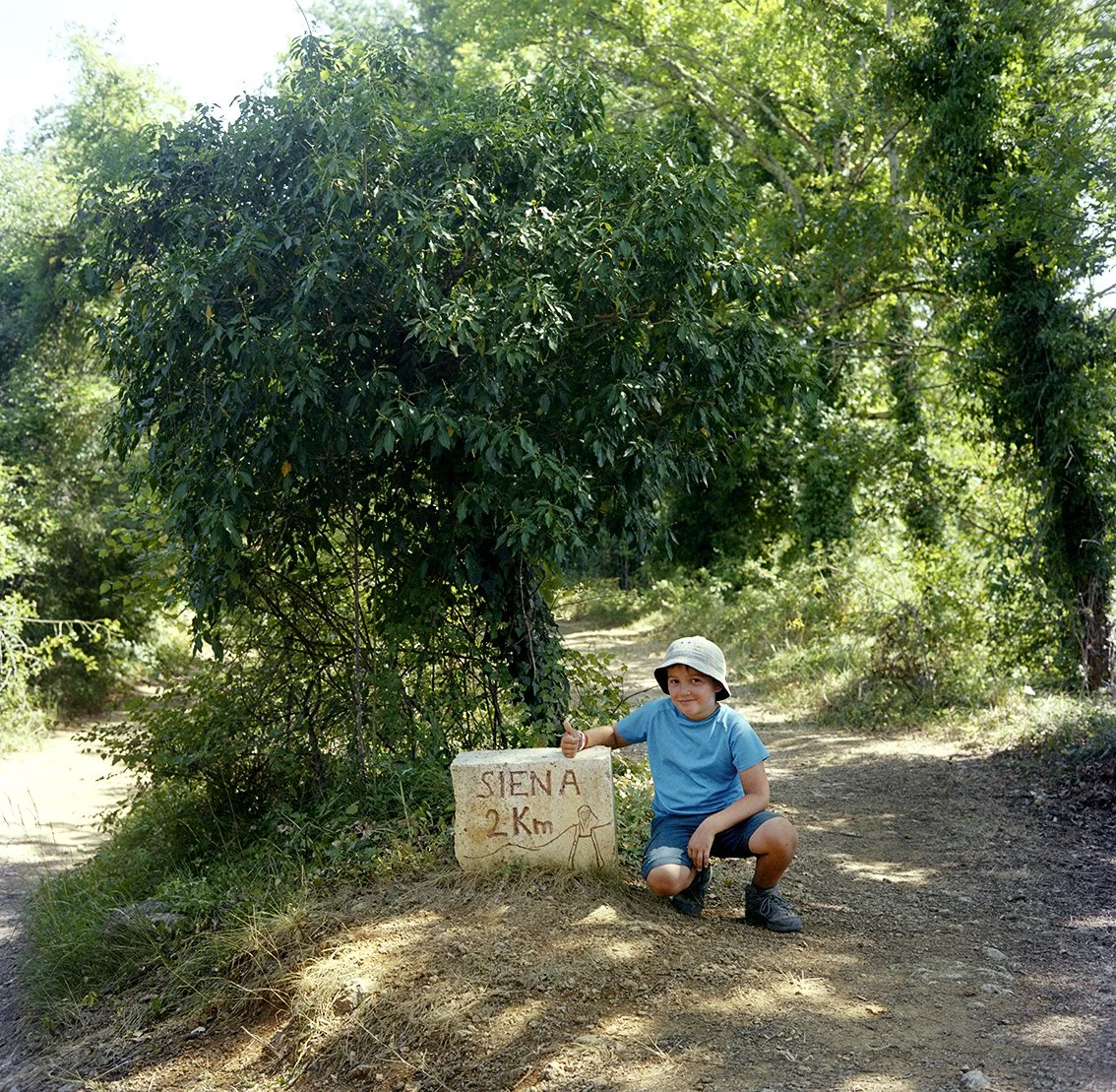 A young boy in hiking gear, wearing a hat, poses next to a sign reading "SIENA 2 km" in a green forest.