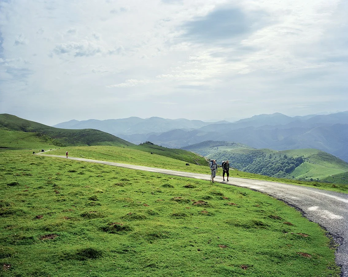 Two hikers walk along a path through a landscape of green hills, with distant mountains under a cloudy sky.
