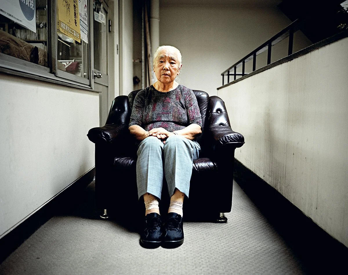 Une femme âgée assise sur un fauteuil en cuir noir dans un couloir étroit, avec un mur blanc à droite et une vitrine à gauche. Elle porte un haut à motifs et un pantalon gris.
